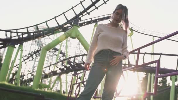 Привлекательная девушка в очках, стоящая возле американских горок при ярком солнечном свете — стоковое видео