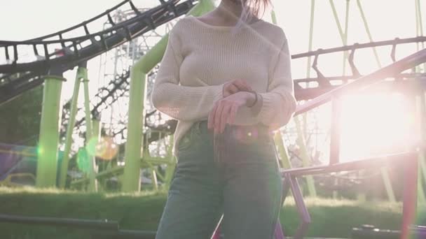 戴着眼镜的漂亮年轻女孩站在明亮的阳光下, 坐在云霄飞车附近 — 图库视频影像