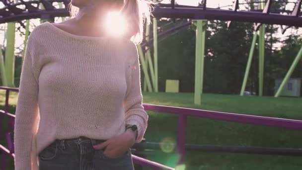 Belle jeune fille dans des lunettes debout près des montagnes russes dans un rayon de soleil lumineux — Video