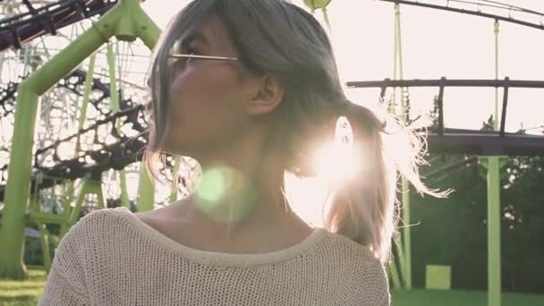 Sexy jonge meisje in glazen staande in de buurt van de achtbaan in de felle zon balk — Stockvideo