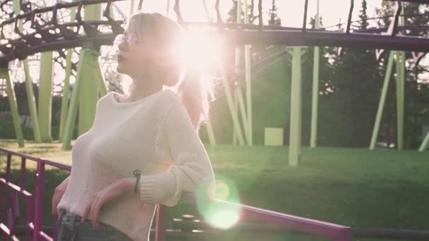 Сексуальная молодая девушка в очках, стоящая возле американских горок в ярком солнечном луче — стоковое видео