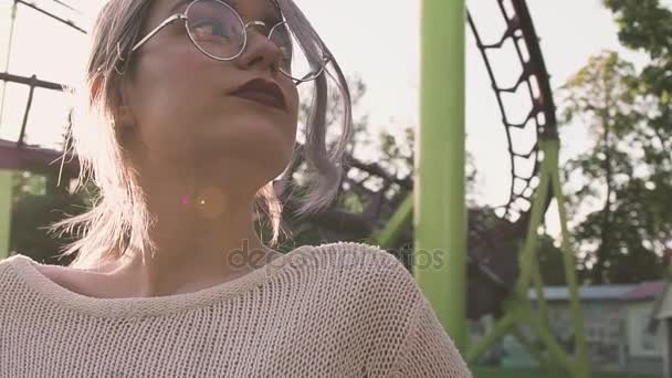 Hübsches junges Mädchen in Brille posiert in der Nähe von Achterbahn im grellen Sonnenlicht — Stockvideo