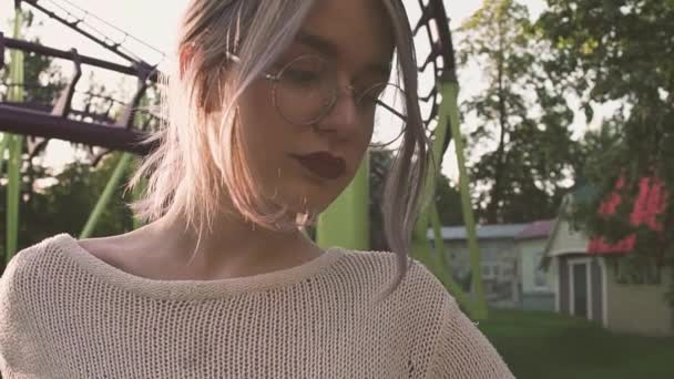 Pretty young girl in glasses posing near roller coaster in bright sun ray — Stock Video