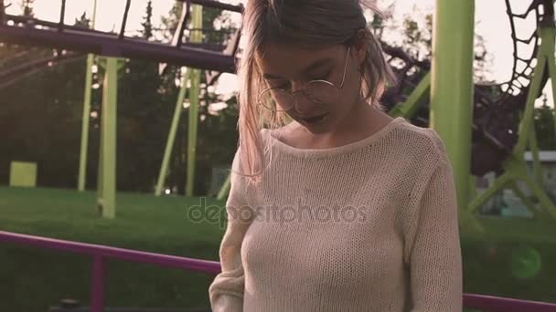 Ελκυστική νεαρή κοπέλα σε ποτήρια θέτοντας κοντά σε τρενάκι του λούνα παρκ στο λαμπρό ήλιο ray — Αρχείο Βίντεο