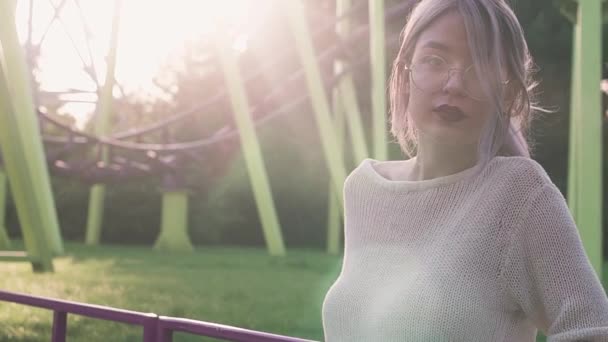 Mooi jong meisje in glazen poseren in de buurt van de achtbaan in de felle zon balk — Stockvideo