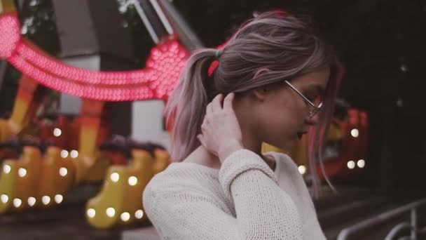 Hipster chica joven en gafas acariciar el pelo cerca de swing atracción en parque de atracciones — Vídeos de Stock