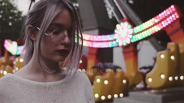 Хипстерская девушка в очках ласкает волосы возле качелей аттракциона в парке развлечений — стоковое видео