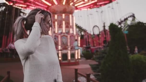 Mooi meisje in glazen strelen haar in de buurt van carrousel attractie in amusment park — Stockvideo