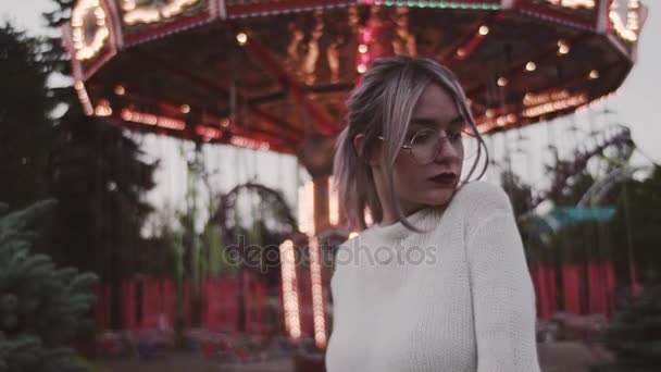 Elegante chica joven en gafas de pie cerca de atracción carrusel en el parque de atracciones — Vídeos de Stock