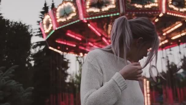 Hezká dívka v brýlích se třese vlasy poblíž objezdu atrakcí v amusment parku — Stock video