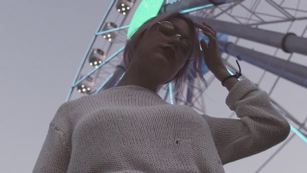 Beautiful young girl in glasses posing under ferris wheel with flashing lights — Stock Video