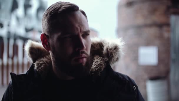 Молодой бородатый мужчина в зимней куртке смотрит в камеру в городской зоне — стоковое видео