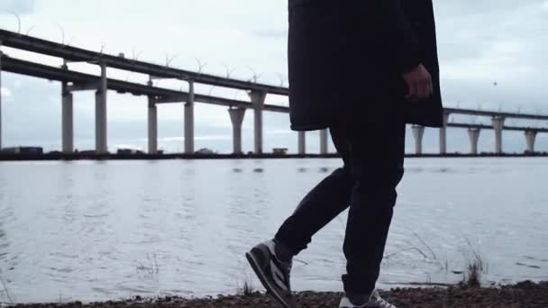 Person in stylischer Winterkleidung spaziert am felsigen Ufer mit Brücke Autobahn — Stockvideo