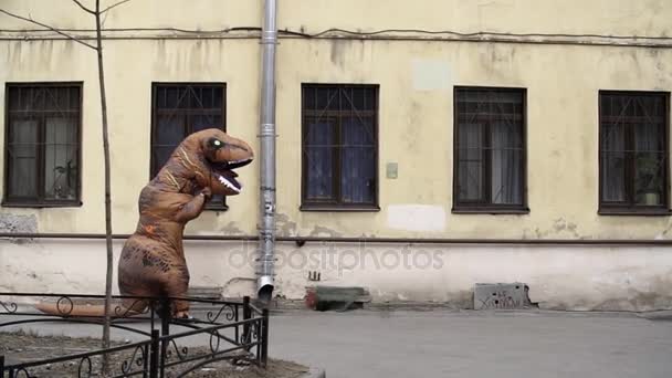 Persona con traje de mascota t rex camina a lo largo de las ventanas en la casa de la ciudad — Vídeo de stock