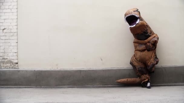 Persoon in t rex mascotte kostuum leunt op de witte muur en loopt af — Stockvideo