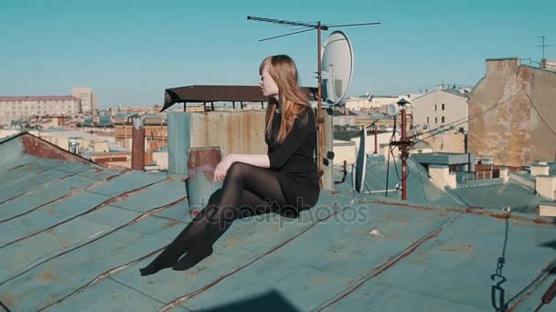 Muchacha joven atractiva en vestido negro sentado en el techo con paisaje urbano escénico — Vídeos de Stock