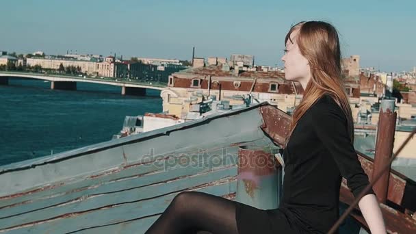 Hermosa mujer joven en vestido negro sentado en el techo con paisaje urbano escénico — Vídeos de Stock