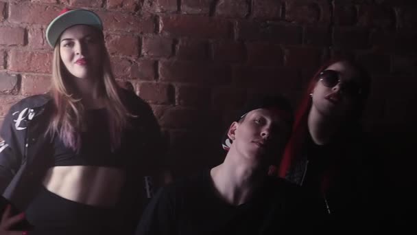 Молодой хип-хоп человек в кепке с татуировками на лице перед кирпичной стеной с девушками — стоковое видео