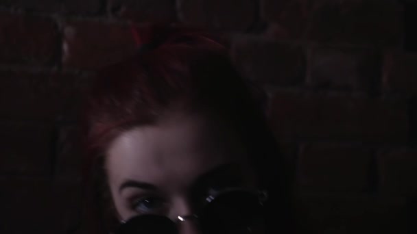 Chica pelirroja en gafas de sol redondas posando frente a la pared de ladrillo en habitación oscura — Vídeo de stock