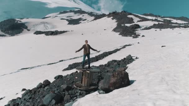 Стрелок с воздуха раскинул руки стоя на кабельной катушке по камням в снежных горах — стоковое видео