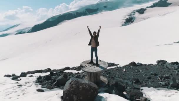 Letecký snímek muž jásající stojící na kabelové cívky na kamenech na zasněžené hory — Stock video