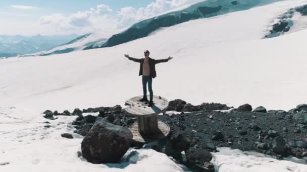 Εναέρια shot άνθρωπος υψώνεται όπλων που στέκεται πάνω σε καρούλι καλωδίου σε πέτρες σε χιονισμένα βουνά — Αρχείο Βίντεο