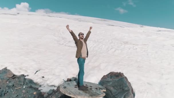 Vue aérienne homme lève les bras debout sur la bobine de câble sur les pierres dans les montagnes enneigées — Video