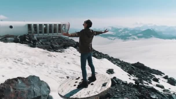 Vue aérienne homme lève les bras debout sur la bobine de câble sur les pierres dans les montagnes enneigées — Video