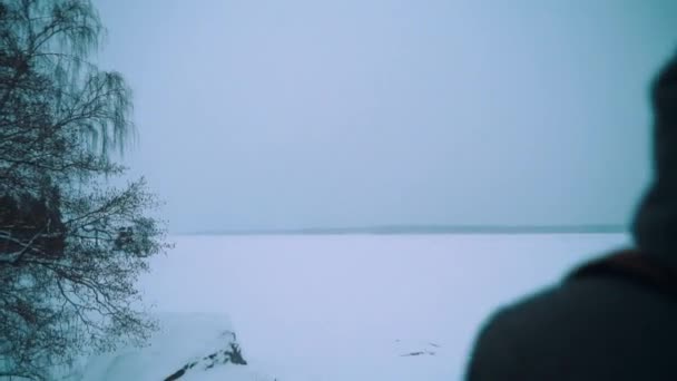 Viaggiatore uomo con zaino in piedi sulla riva del lago innevata nella giornata invernale — Video Stock