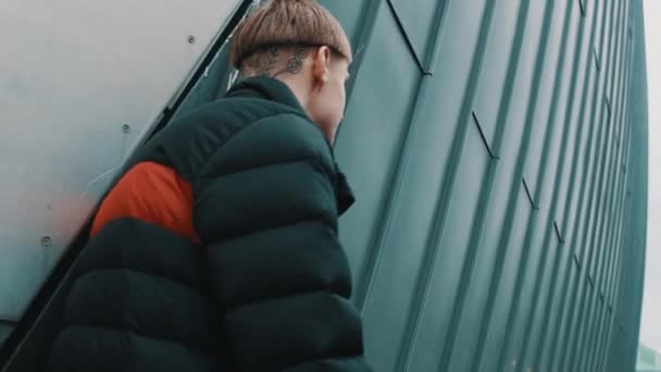 Joven chico con estilo con tatuajes sentado cerca de la pared de metal — Vídeo de stock