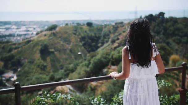 Ung kvinna i vit klänning står på terrassen i villa tittar på staden — Stockvideo