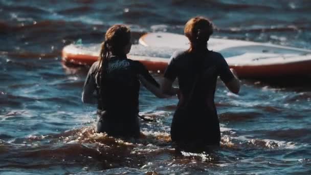 Dua gadis berpakaian renang berjalan dalam air bergelombang menuju papan selancar mengambang — Stok Video