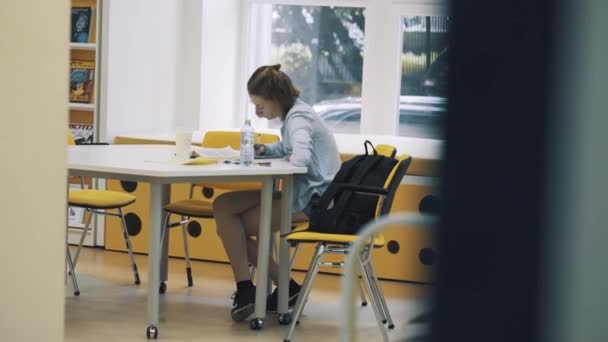 Jeune jolie fille étudie à la bibliothèque assis jambe croisée à la table — Video