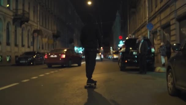 Скейтбордист хлопець у чорний балахон їде на проспекті міста ніч — стокове відео