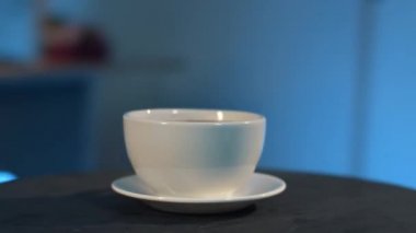 Kamera beyaz fincanın etrafında dönüyor ve siyah masa üzerinde fincan tabağı, mavi arkaplan.