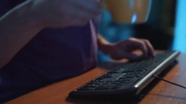 Kamera, siyah klavyede yazan mor tişörtlü adamın elleri etrafında dönüyor.