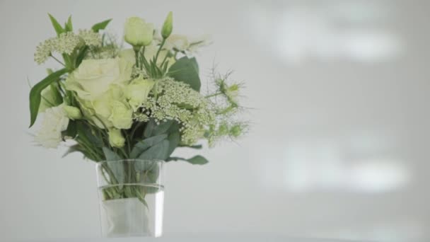 Lindas flores blancas con grandes hojas verdes en jarrón de vidrio puesto en la mesa blanca . — Vídeo de stock