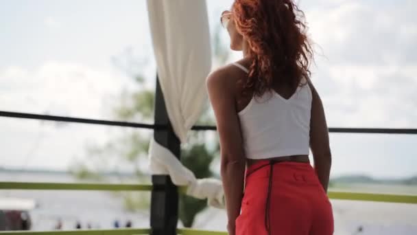 MOSCÚ, RUSIA - 12 de julio de 2019: Mujer joven con el pelo rizado rojo en camiseta blanca y pantalones cortos baila en el resort — Vídeos de Stock