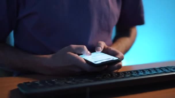 Mince gars en t-shirt assis au bureau en face du clavier et tapant sur le smartphone .
