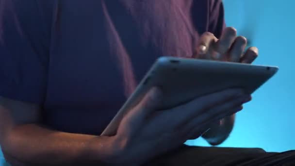 Hombre joven en camiseta violeta se sienta en habitación oscura y activamente usando su tableta . — Vídeos de Stock