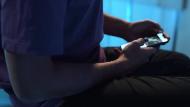 Hombre en camiseta violeta se sienta solo en habitación oscura y mira a través del teléfono celular — Vídeos de Stock