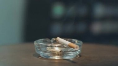 Kamera, kenarında yanan sigarayla cam kül tablasının etrafında döner..