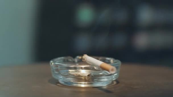 Camera rotates around glass ashtray with burning cigarette placed on its edge. — Stock Video