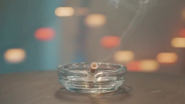 Kamera obraca się wokół szklanej popielniczki z płonącym papierosem umieszczonym na krawędzi. — Wideo stockowe