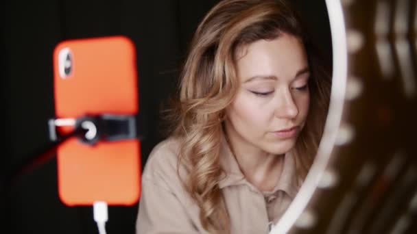 Женщина-видеоблогер использует подводку для глаз, держа зеркало в руке перед кольцом света — стоковое видео