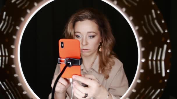 Mature blonde female beauty blogger holds orange smartphone and taps it. — Stock Video