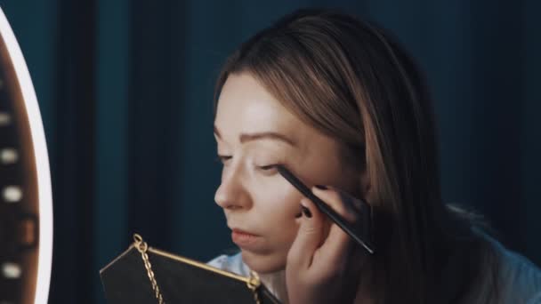 Woman beauty vlogger uses eyeliner holding mirror in hand in front of ring light — Stock Video