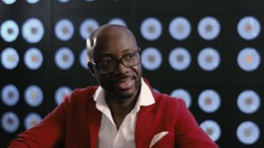 Kırmızı takım elbiseli, beyaz gömlekli ve şık gözlüklü gülümseyen kel, olgun Afro-Amerikan işadamı bilgisayarda çalışıyor, bulanık arka planı olan karanlık bir odada oturuyor ve mavi klasik bir renge sahip..