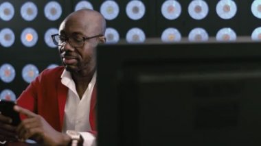 Siyah sakallı, olgun bir iş adamı, kırmızı takım elbiseli, beyaz gömlekli ve şık gözlükleri olan modern akıllı telefona bakıyor, elinde tutuyor, koyu renk arka planda mavi renkli bir ofiste oturuyor..