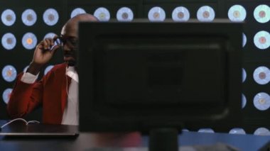 Kırmızı takım elbise, beyaz gömlek, şık gözlükler ve beyaz telefon kulaklığı takan Afro-Amerikan sakallı adam bilgisayar başında müzik dinliyor, karanlık odada klasik arka planda oturuyor..